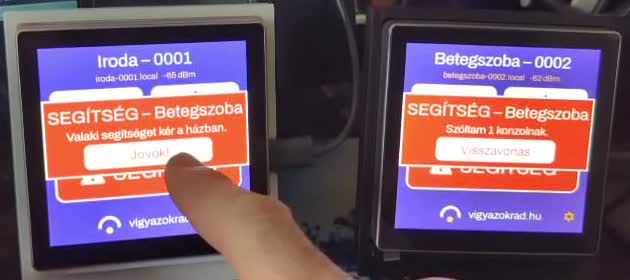 Ugyanaz a két konzol riasztás közben. A hívó panelen „Szóltam 1 konzolnak” és egy Visszavonás gomb; a másikon piros sávban „SEGÍTSÉG — Betegszoba, valaki segítséget kér a házban”, és egy ujj épp megnyomja a „Jövök!” gombot.